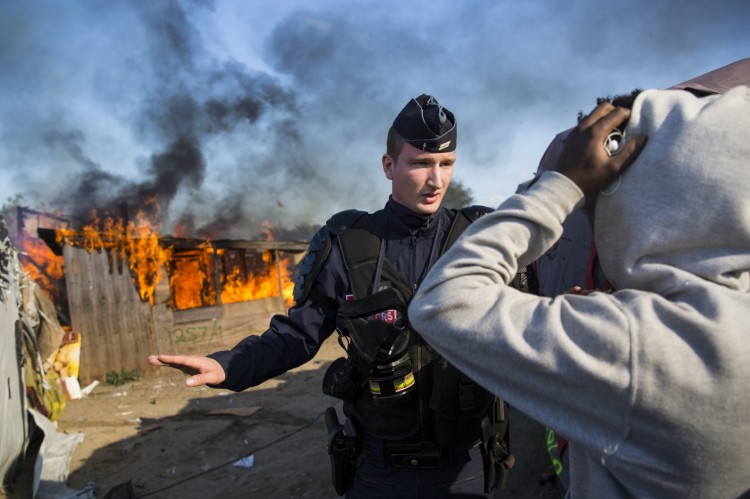 A police officer controls the crowd Monday while behind him officials set fire to “The Jungle” refugee camp at Calais, France, where about 8000 Afghan and African refugees, many of them unaccompanied children, have lived while they hoped to get asylum in Britain. British civilians, not the French, provided food, clothes and social services to the migrants; some were more than ready to leave, but others will miss the little bit of home they were able to create. Most of the refugees are being dispersed throughout the French countryside, often to towns where people don’t want them and are greeting them with jeers and taunts. (Jack Taylor/Getty Images)
